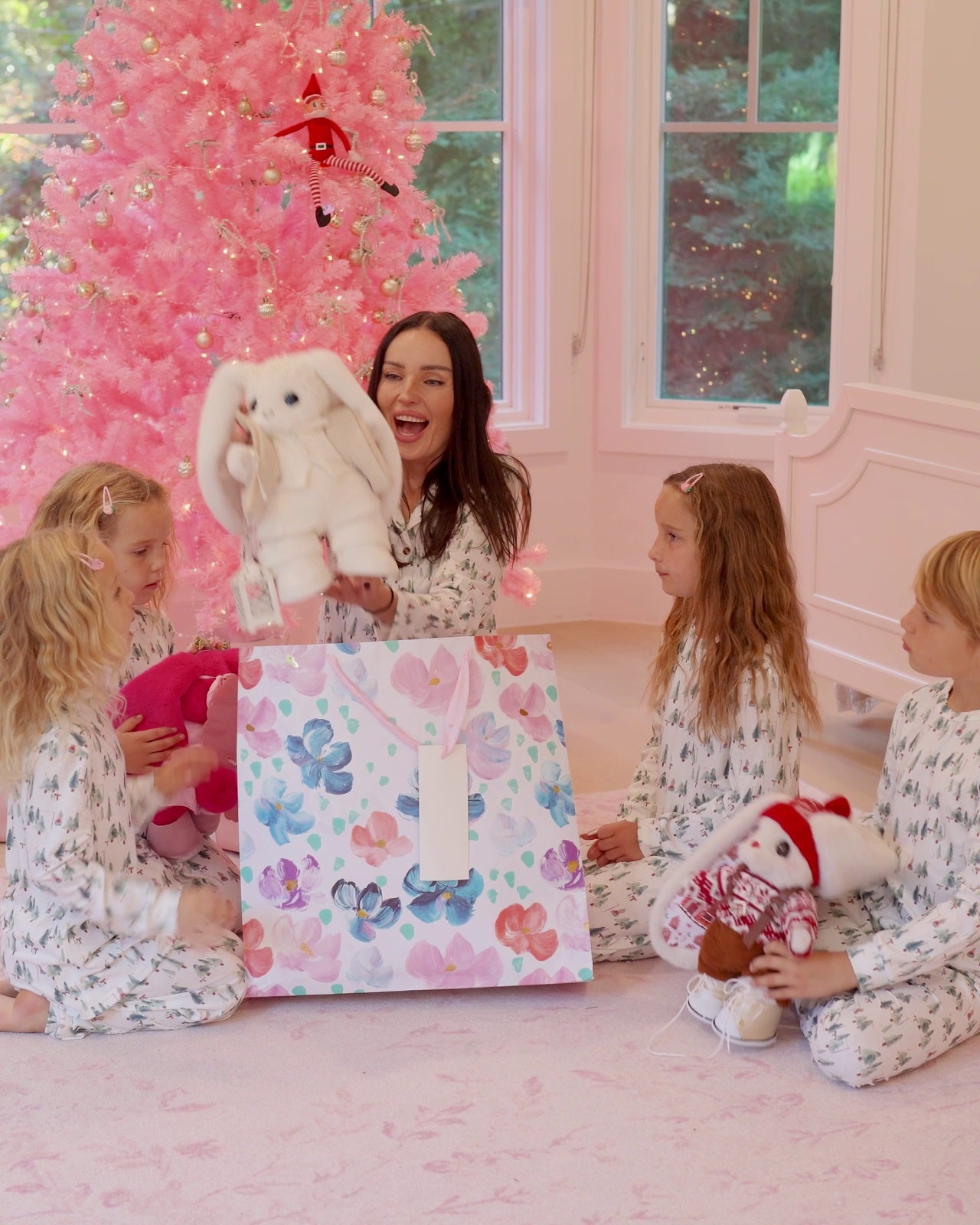 A woman and four children sitting on a pink floor in front of a pink Christmas tree, gathered around a large floral gift bag with a white number 1 card. One child holds a white plush bunny, and another holds a small doll wearing a red Santa hat.