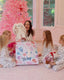 A woman and four children sitting on a pink floor in front of a pink Christmas tree, gathered around a large floral gift bag with a white number 1 card. One child holds a white plush bunny, and another holds a small doll wearing a red Santa hat.