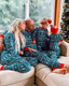 A family of four wearing matching teal and white Christmas-themed patterned pajamas, sitting together on a beige couch in front of a window with a decorated Christmas tree visible in the background.